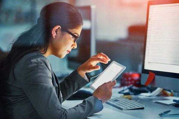 Female IT professional using tablet at night next to a desktop computer screen