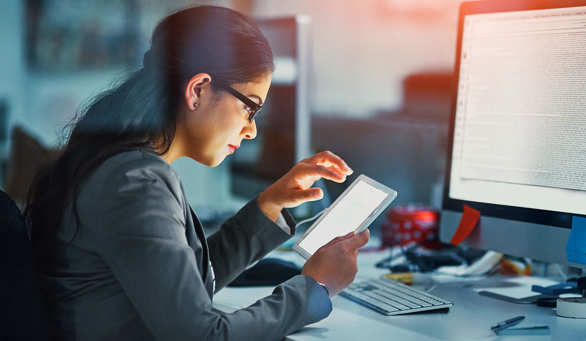 Female IT professional using tablet at night next to a desktop computer screen